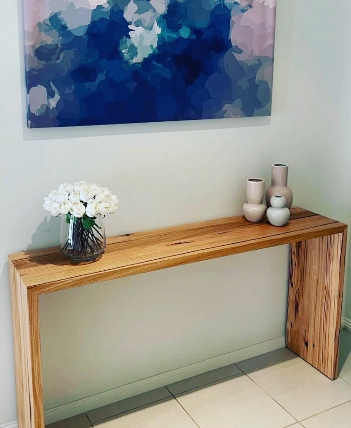 A wooden console table with a steel base, featuring a waterfall design, placed against a wall with a floral arrangement and decorative vases on top.